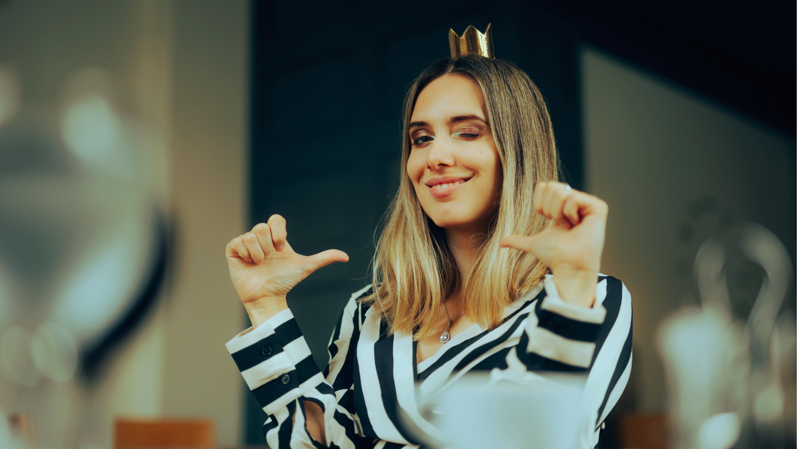 mulher olhando para quem vê a foto, piscando com um dos olhos, usando uma pequena coroa dourada, aponta para si mesma com ambos os dedões das mãos, com um sorriso malicioso, expressando autoconfiança